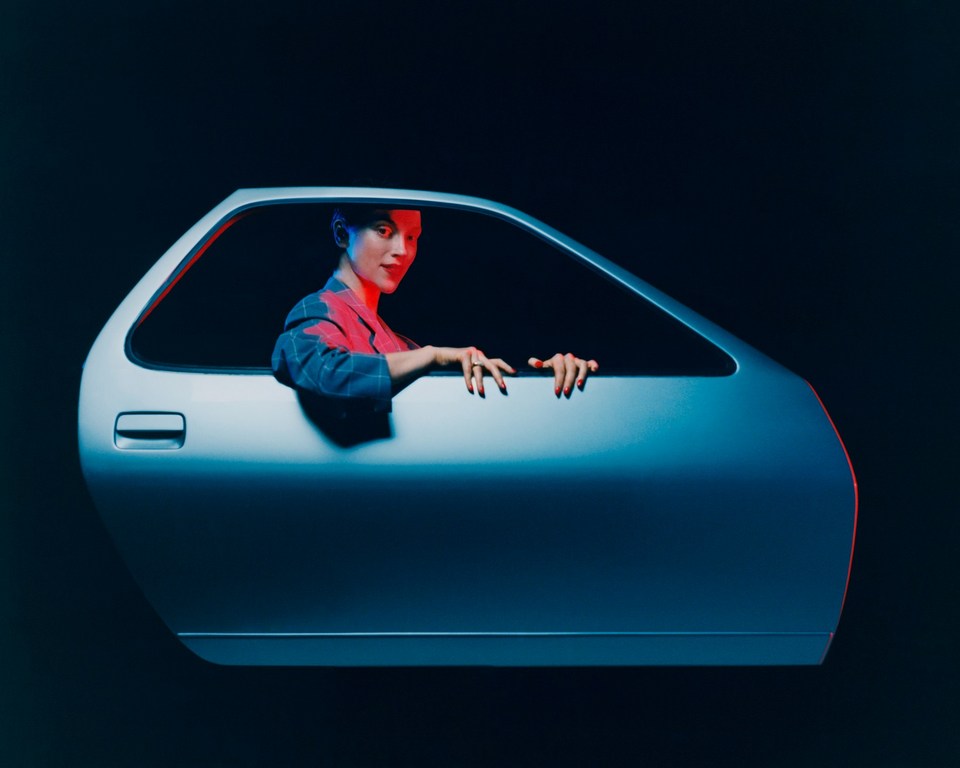 Woman poses framed by a car door against a dark background, lit with blue and red studio lights. Automotive and fashion photography.