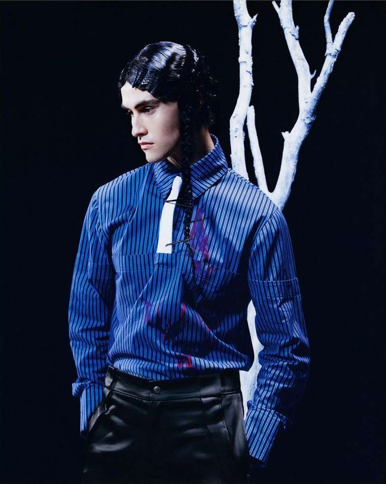 Portrait of a young adult male model in a striped shirt and black pants standing in front of a white branch against a dark backdrop.