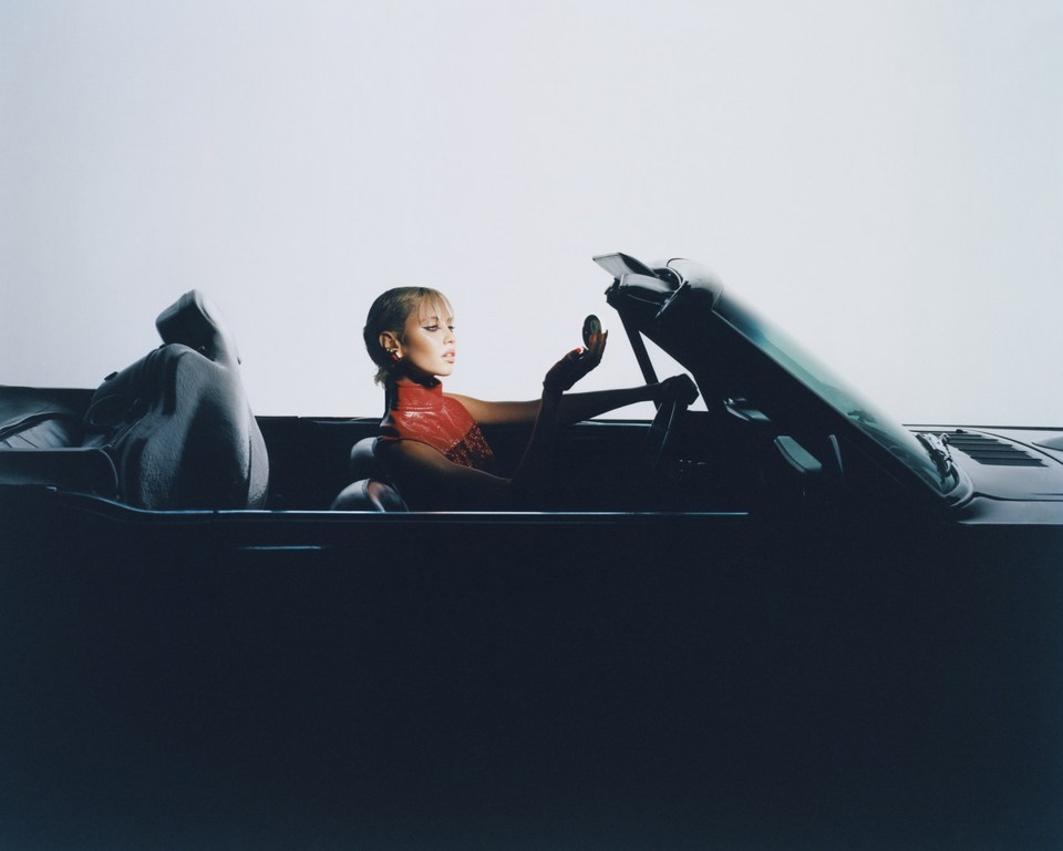 Woman in red outfit sits in a dark convertible car looking in a mirror. Plain white background. Automotive and beauty theme.