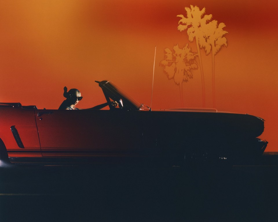 A red convertible car with a person driving against an orange gradient background with palm tree silhouettes, evokes a retro vibe.