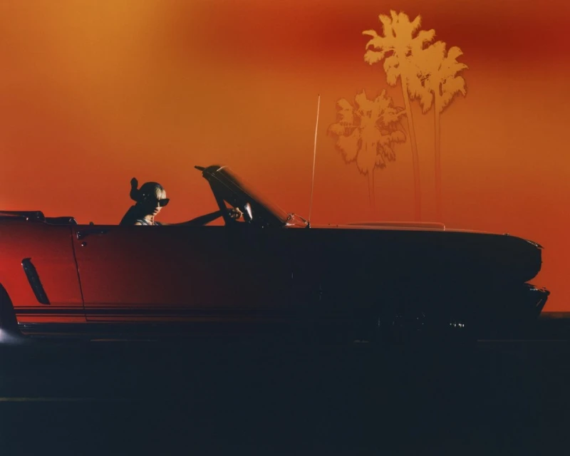 A red convertible car with a person driving against an orange gradient background with palm tree silhouettes, evokes a retro vibe.