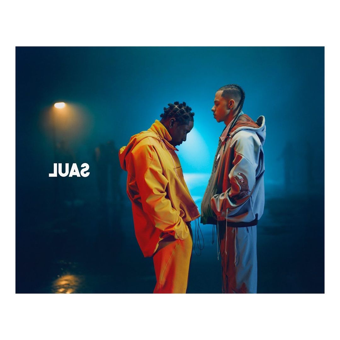 Two hip-hop artists pose indoors in matching streetwear against a blue background with soft lighting and visible text 'JUAS'.