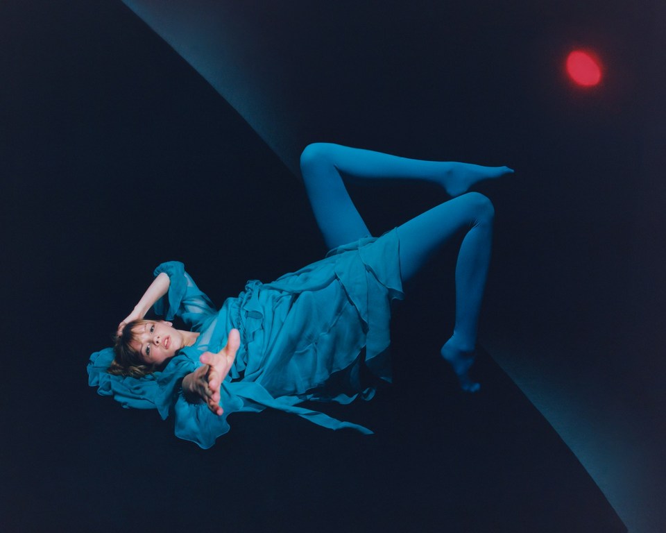 A young woman in blue tights and dress is lying on a black surface, arm outstretched, with a soft red light illuminating the scene.