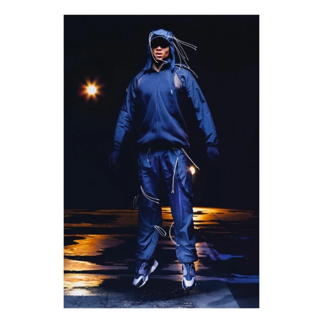 Full-length studio shot of adult male wearing blue hooded tracksuit and sneakers, jumping with a dark background and reflection on the floor.
