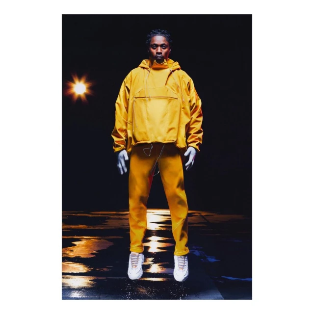 An adult man in a yellow tracksuit, white gloves, and sneakers stands in a dark space illuminated by an off-center light, creating shadows.