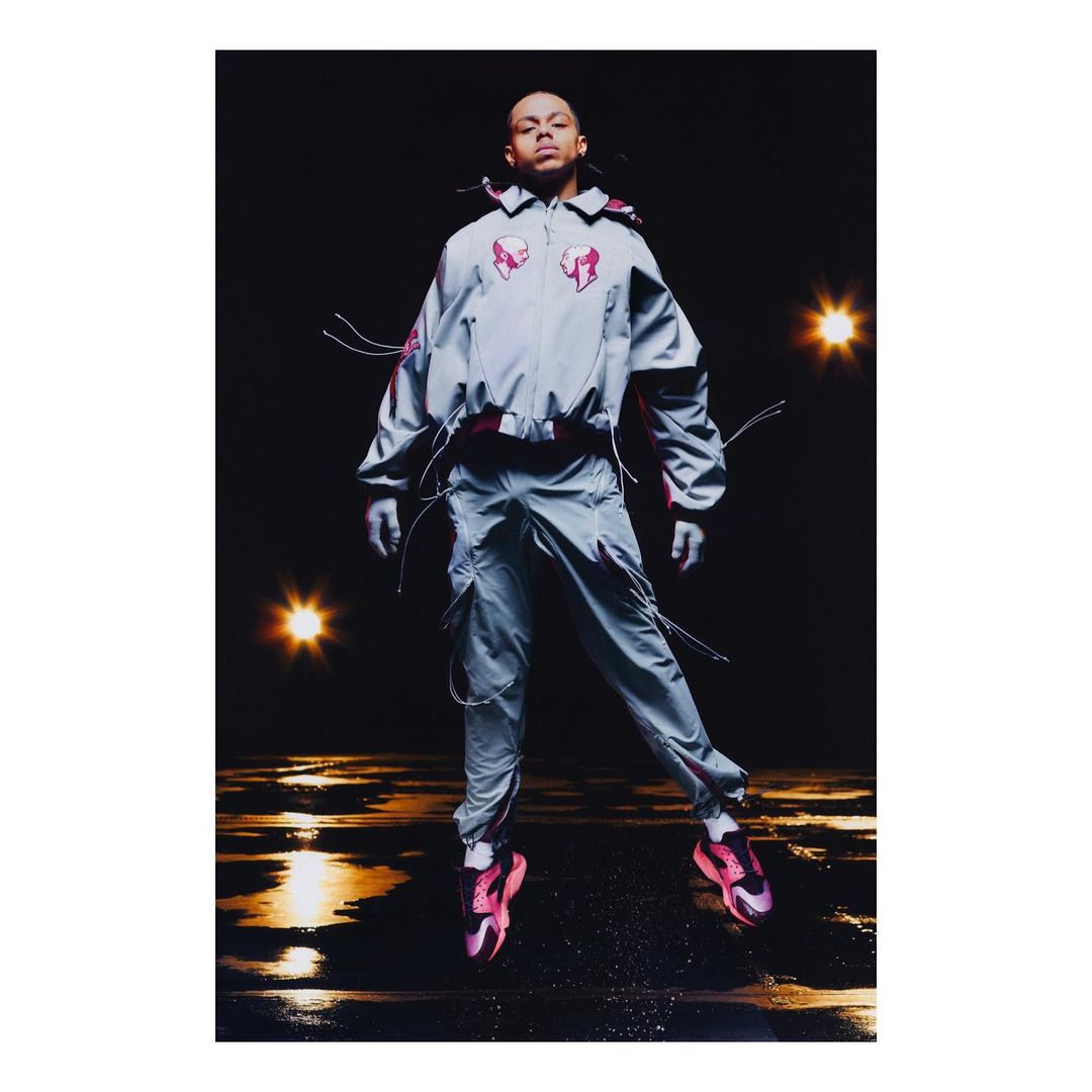 Person in grey fashion tracksuit and pink sneakers jumping in air, shot at eye-level in photo studio with dark background.
