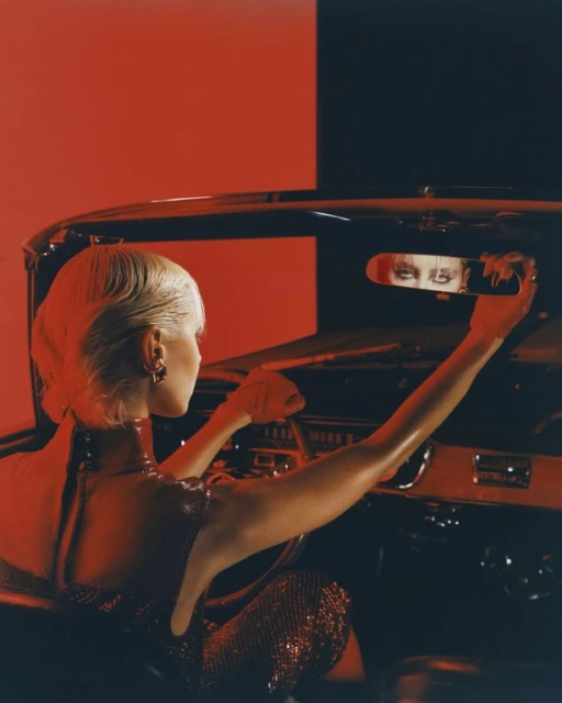 Woman in a car adjusting the rearview mirror, wearing a sparkling red dress and short blonde hair, bathed in saturated red lighting.