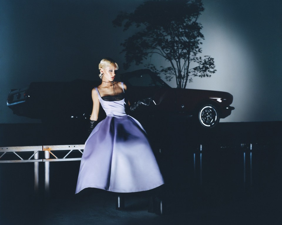 Blonde woman poses in a lavender dress with a black bodice, gloves and necklace in front of a classic car and tree silhouette.