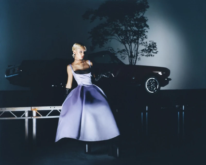 Blonde woman poses in a lavender dress with a black bodice, gloves and necklace in front of a classic car and tree silhouette.