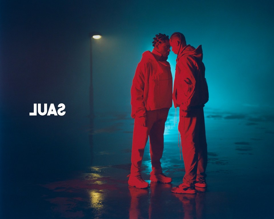 Two people in matching red outfits stand face-to-face under a street light in a gritty urban environment with a teal and blue color palette.