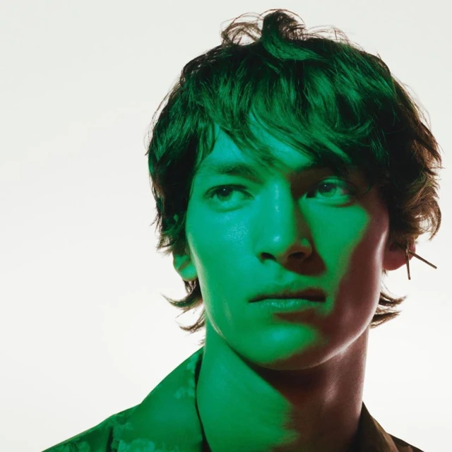 Headshot of a fair-skinned adult male with dark, wavy hair, wearing a patterned shirt, lit with green and red hues against a white backdrop.