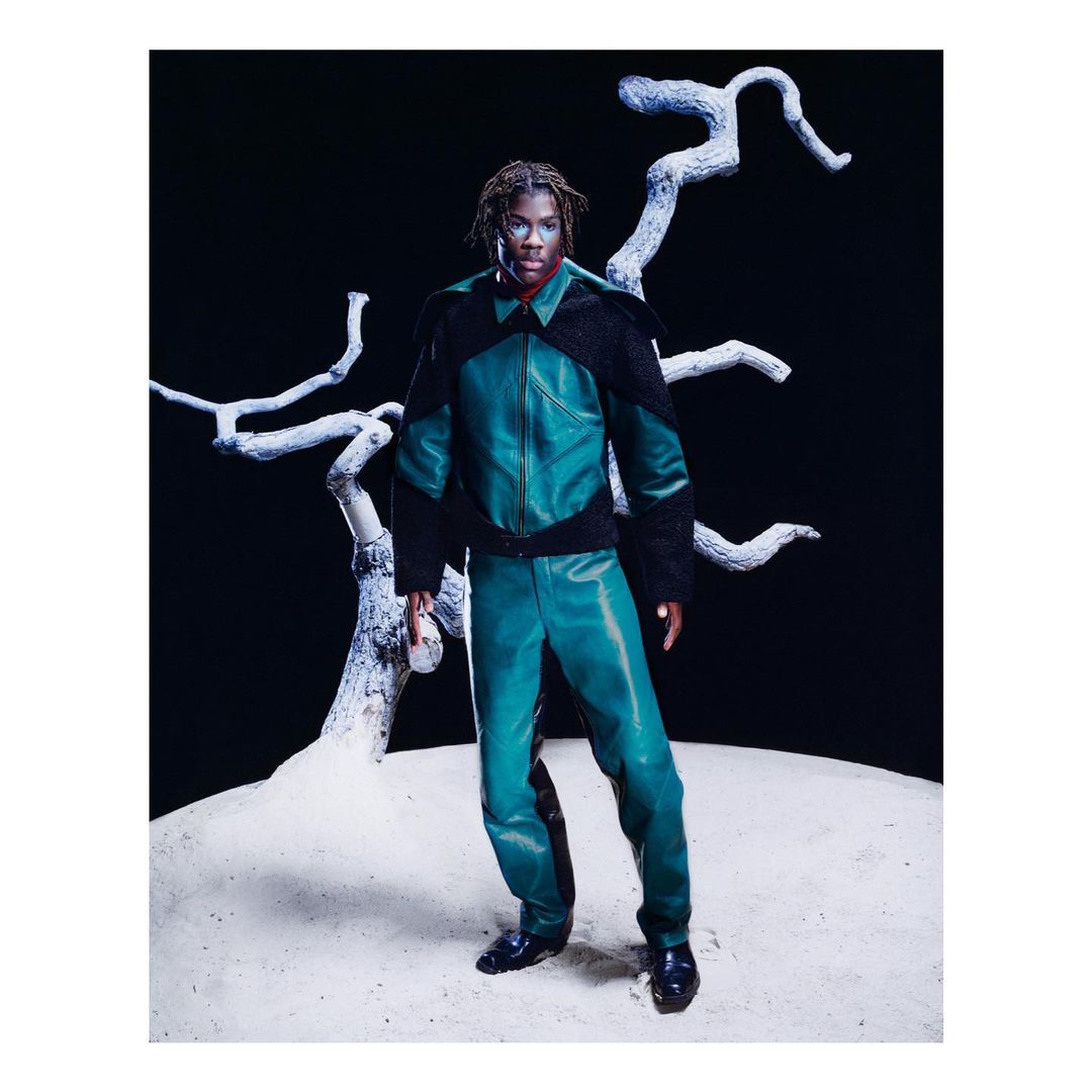 Adult male in fashion shot. Man is wearing a green leather jacket and pants, standing on a sandy surface in front of an abstract white tree.