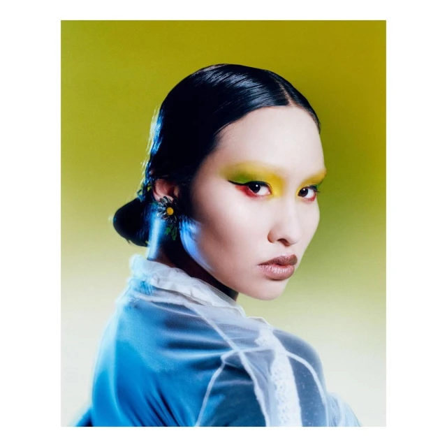 Close-up of an Asian woman with yellow eye makeup and a transparent white top posing against a vibrant yellow-green backdrop.