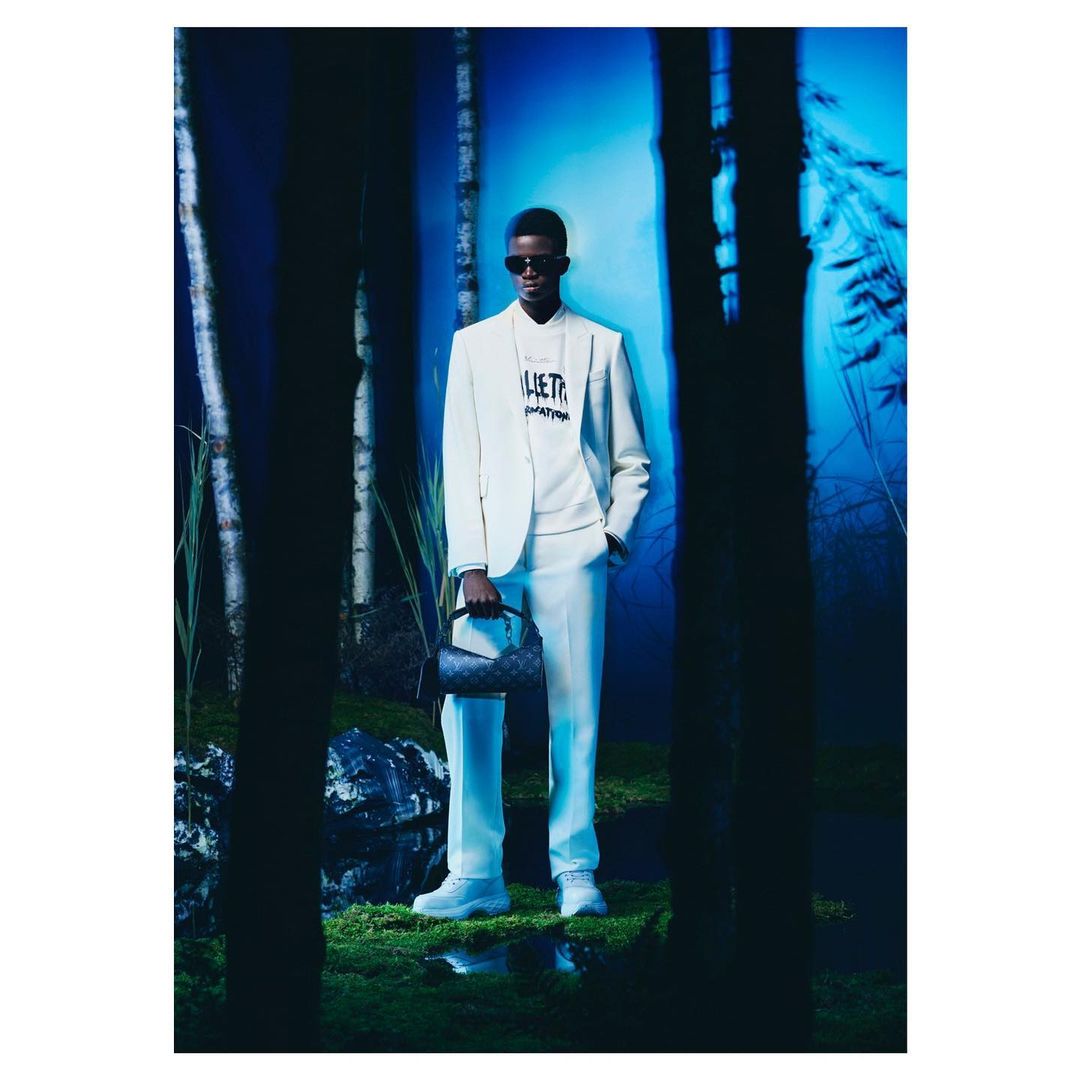 Man poses in an off-white suit in a shadowy outdoor setting with birch trees and a purse; fashion advertisement.