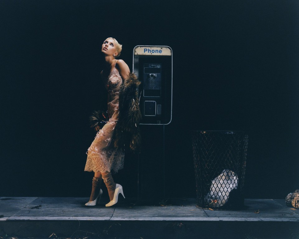 Woman in lace dress and heels stands by a vintage phone booth and trash can in a darkly lit urban setting at night.