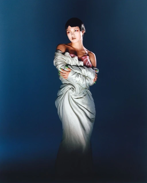 Elegant Asian woman in draped white fabric dress, posing against a gradient blue backdrop in a studio setting, fashion editorial photography.