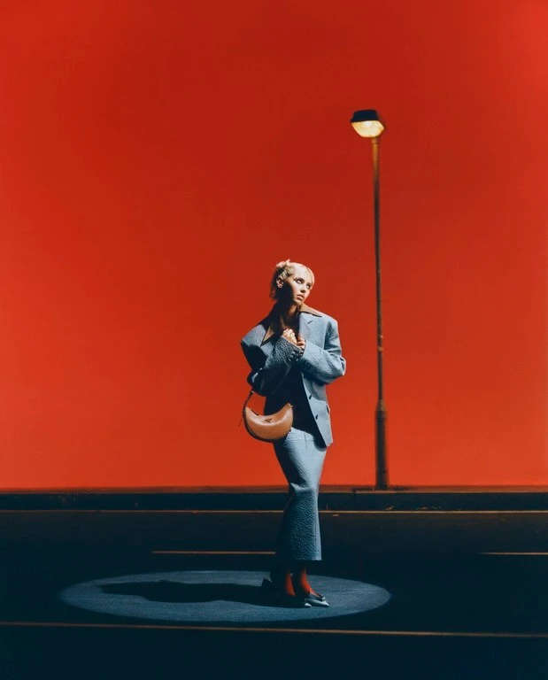 Model in grey blazer and matching skirt standing under a streetlamp against a solid orange background photographed by Scandebergs