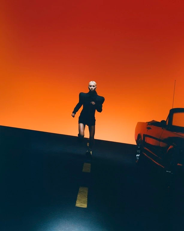 Model in black structured shoulders outfit running on road beside car with orange sunset sky