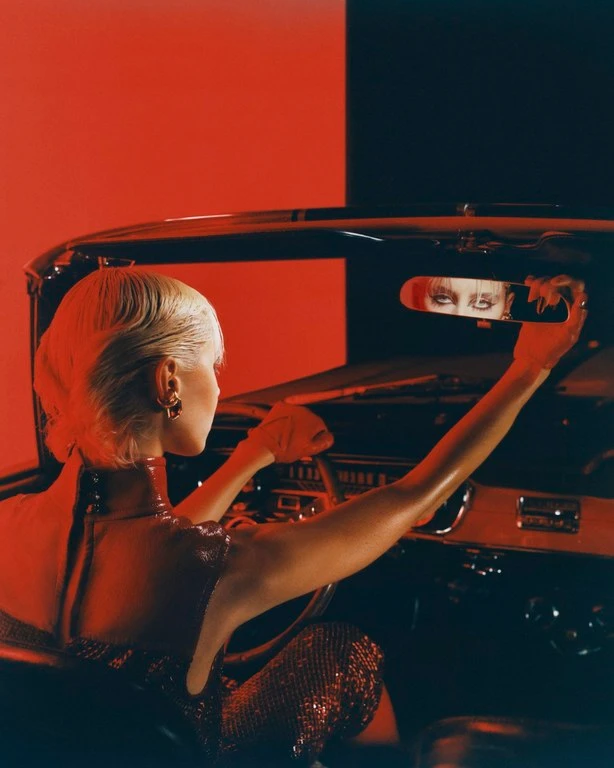 Blonde woman wearing red leather attire inside a vintage car, her eyes reflected in the rearview mirror captured by Scandebergs.