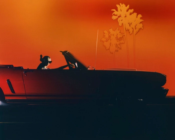 Person in sunglasses driving a red convertible car against a vibrant sunset sky with palm trees by Scandebergs