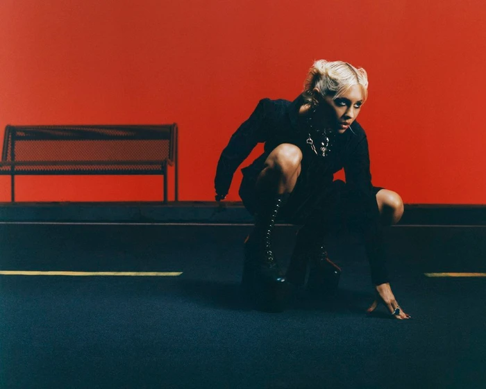 Blonde woman with space buns crouching on asphalt in a black outfit with platform boots against a red wall