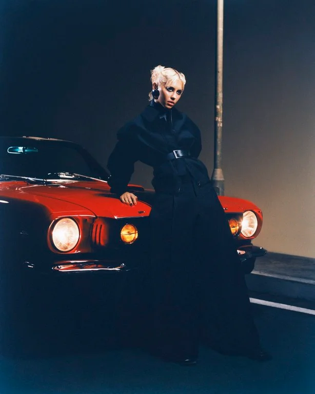 Blonde woman wearing a dark jacket leaning against the hood of a red vintage car at night by Scandebergs