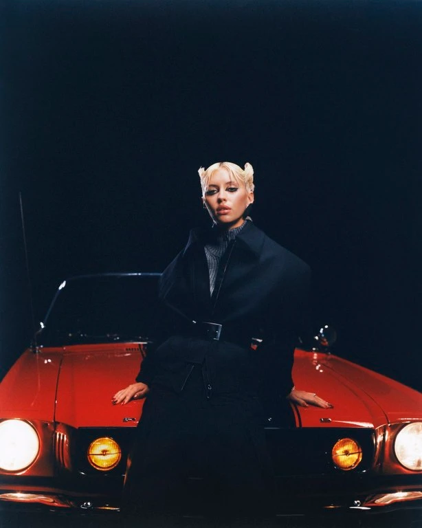 Blonde woman wearing a black jacket leaning against the hood of a red car at night with headlights glowing