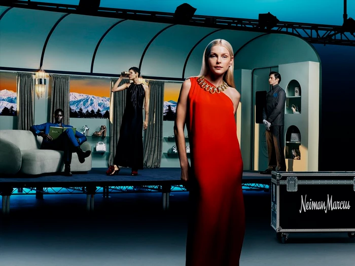 Blonde woman in a bright red dress stands in the foreground of a staged set with several other models and a branded flight case.
