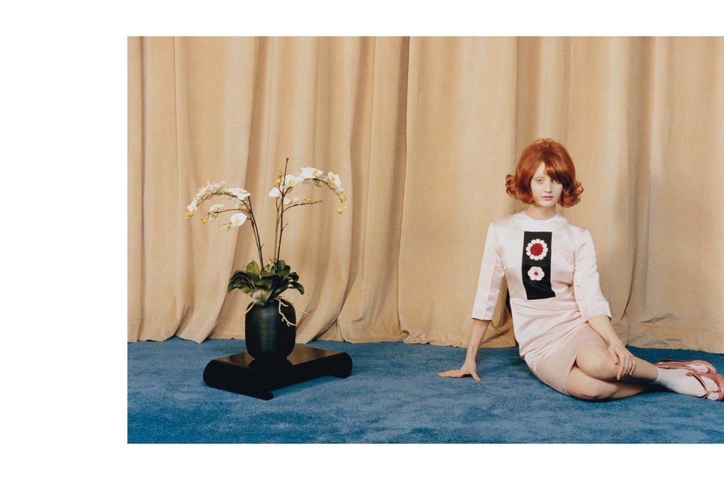 A red-haired woman poses on a blue carpet wearing a light pink dress in front of a beige curtain next to a black pot with white orchids.