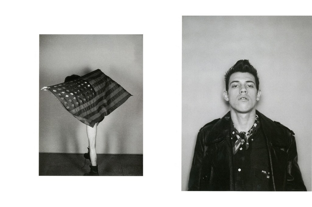 Black and white photo of a man in a leather jacket and a person draped with an American flag standing against a wall.