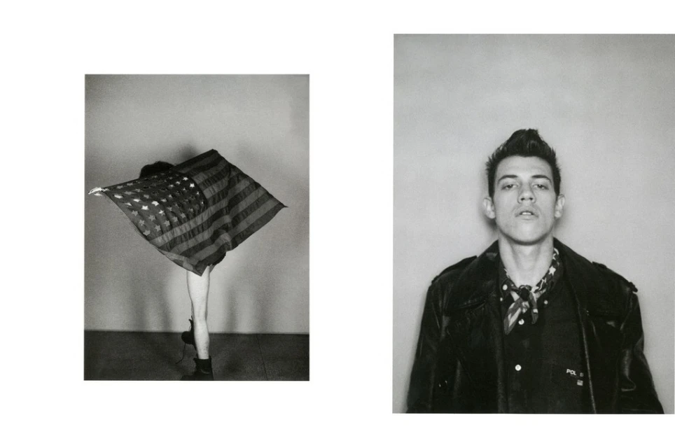 Black and white photo of a man in a leather jacket and a person draped with an American flag standing against a wall.