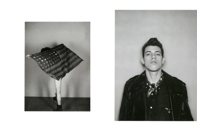 Black and white photo of a man in a leather jacket and a person draped with an American flag standing against a wall.