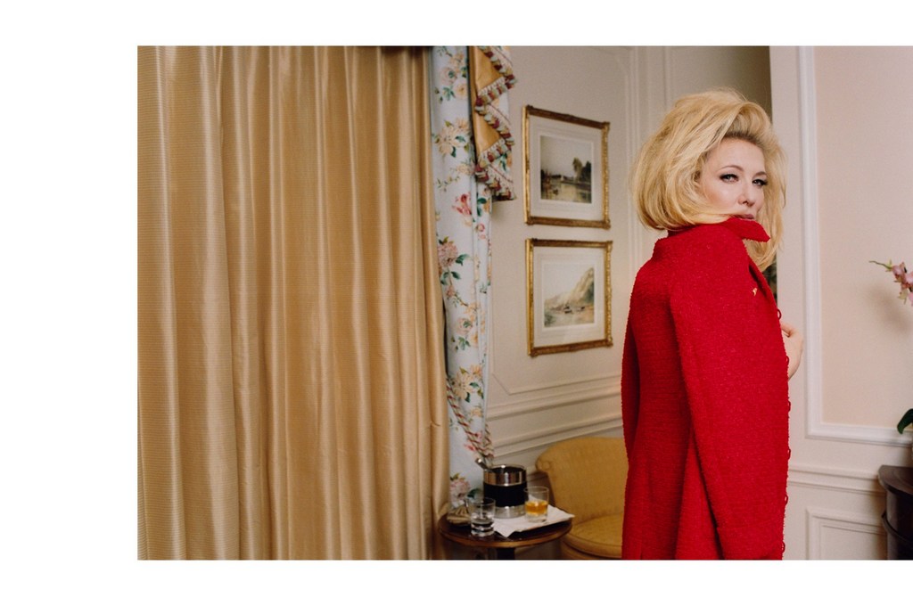 Woman in red coat with blonde hair looks over her shoulder in an indoor setting, with gold curtains and framed artwork in the background.
