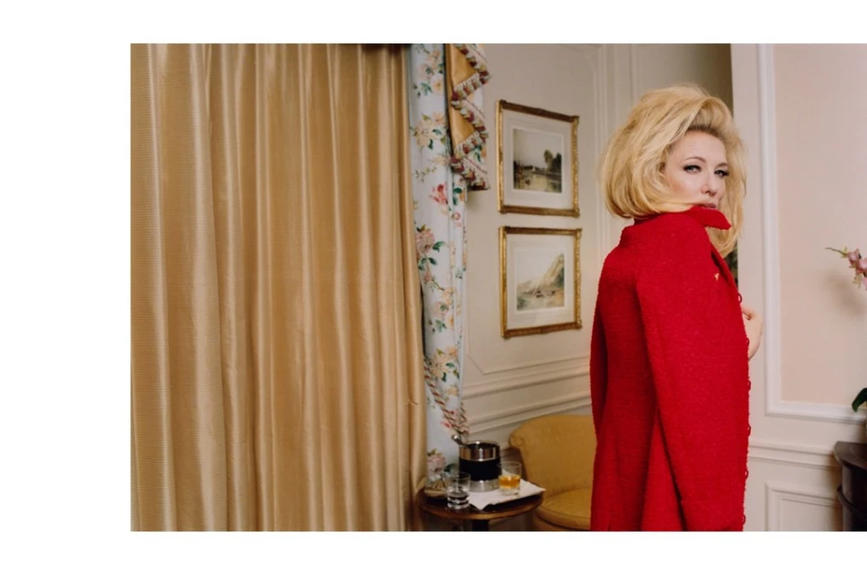Woman in red coat with blonde hair looks over her shoulder in an indoor setting, with gold curtains and framed artwork in the background.
