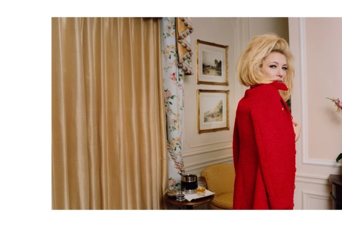Woman in red coat with blonde hair looks over her shoulder in an indoor setting, with gold curtains and framed artwork in the background.