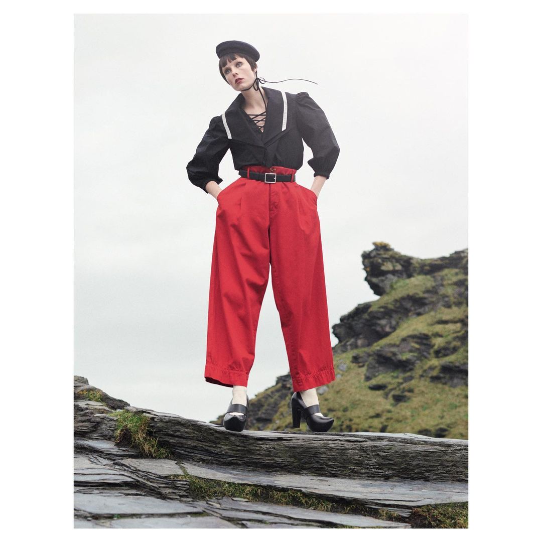 A young woman in a black top and red pants stands on rocks in front of a cloudy sky. She wears a black beret and platform shoes.