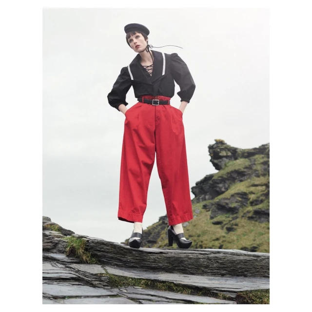 A young woman in a black top and red pants stands on rocks in front of a cloudy sky. She wears a black beret and platform shoes.