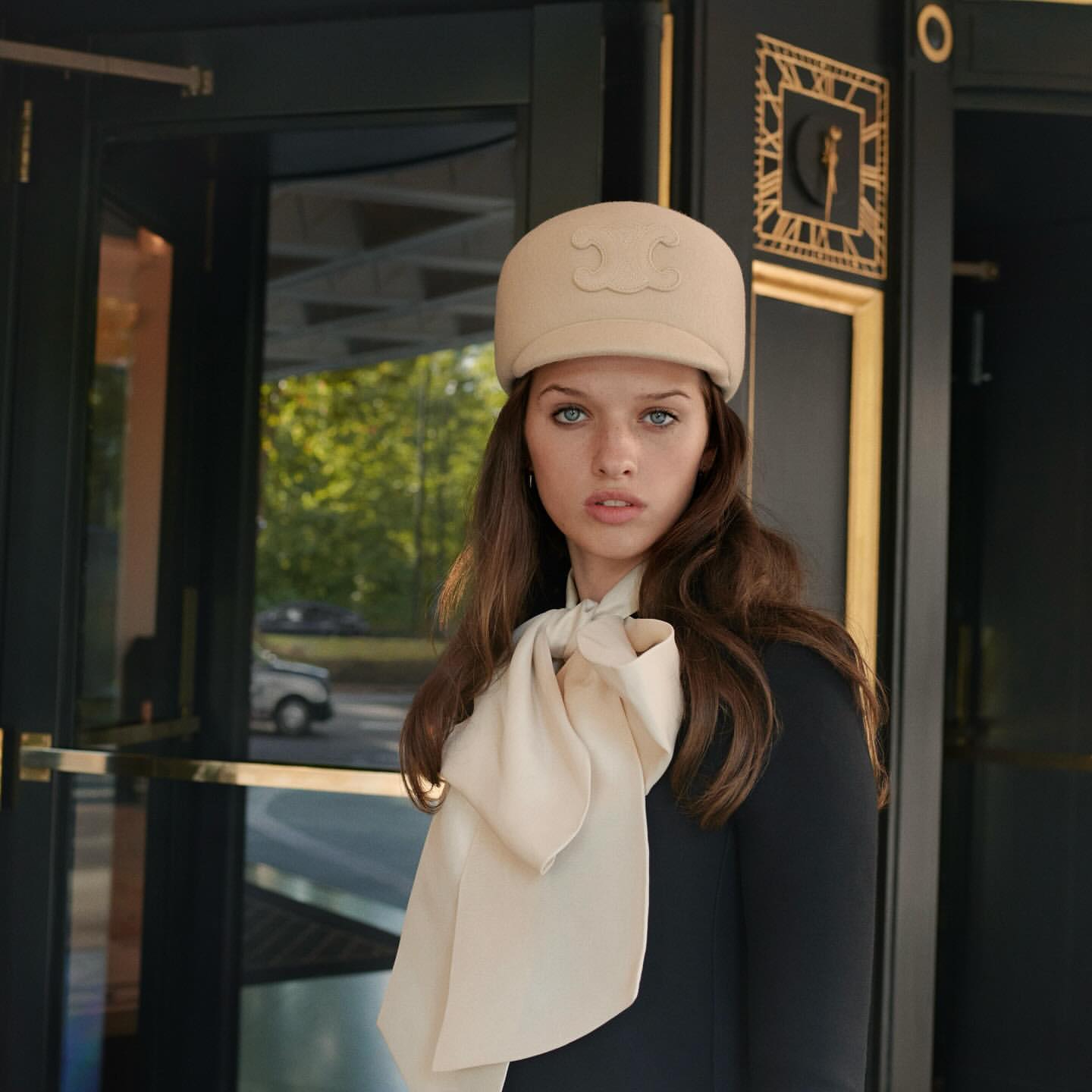 Portrait of a young woman with light hair, wearing a hat and scarf, standing in front of a dark door with green trees behind.