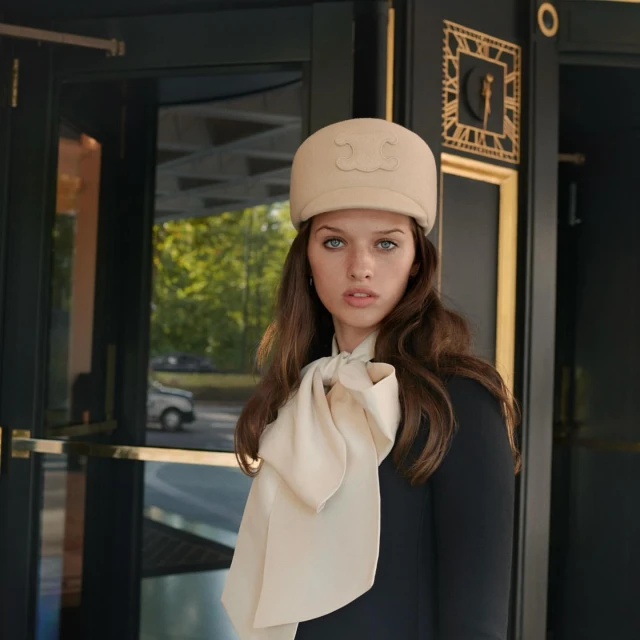 Portrait of a young woman with light hair, wearing a hat and scarf, standing in front of a dark door with green trees behind.