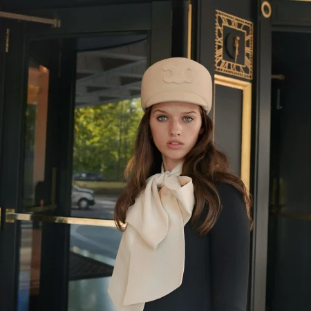 Portrait of a young woman with light hair, wearing a hat and scarf, standing in front of a dark door with green trees behind.