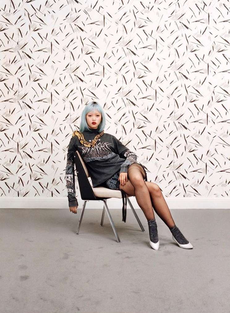 A woman with light blue hair sits on a chair in front of patterned wallpaper, wearing a black long-sleeved shirt and fishnet stockings.
