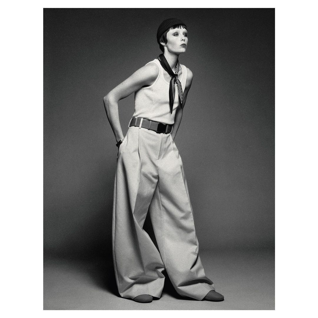 Fashion shot of an adult female model wearing a tank top and baggy pants with a black hat, posing in a photo studio against a grey backdrop.