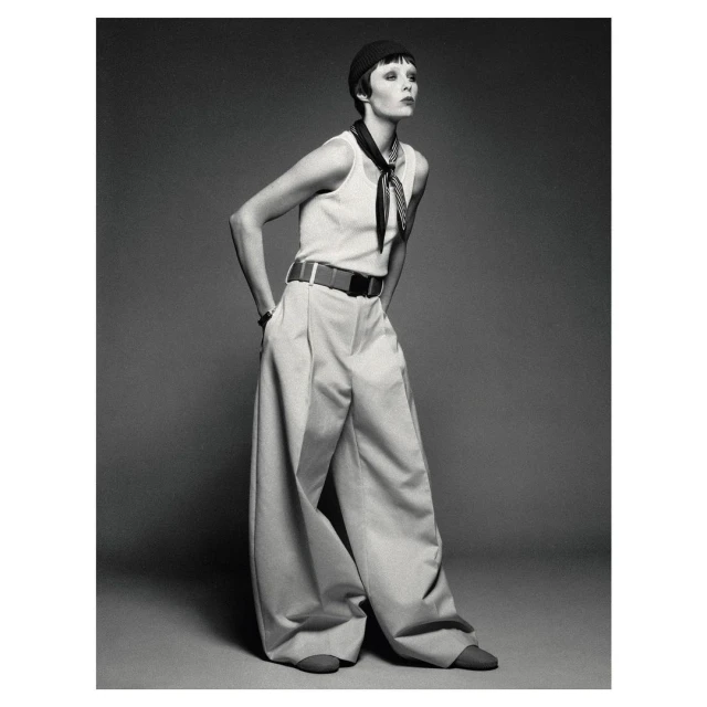 Fashion shot of an adult female model wearing a tank top and baggy pants with a black hat, posing in a photo studio against a grey backdrop.