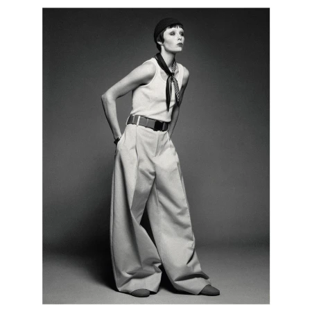Fashion shot of an adult female model wearing a tank top and baggy pants with a black hat, posing in a photo studio against a grey backdrop.