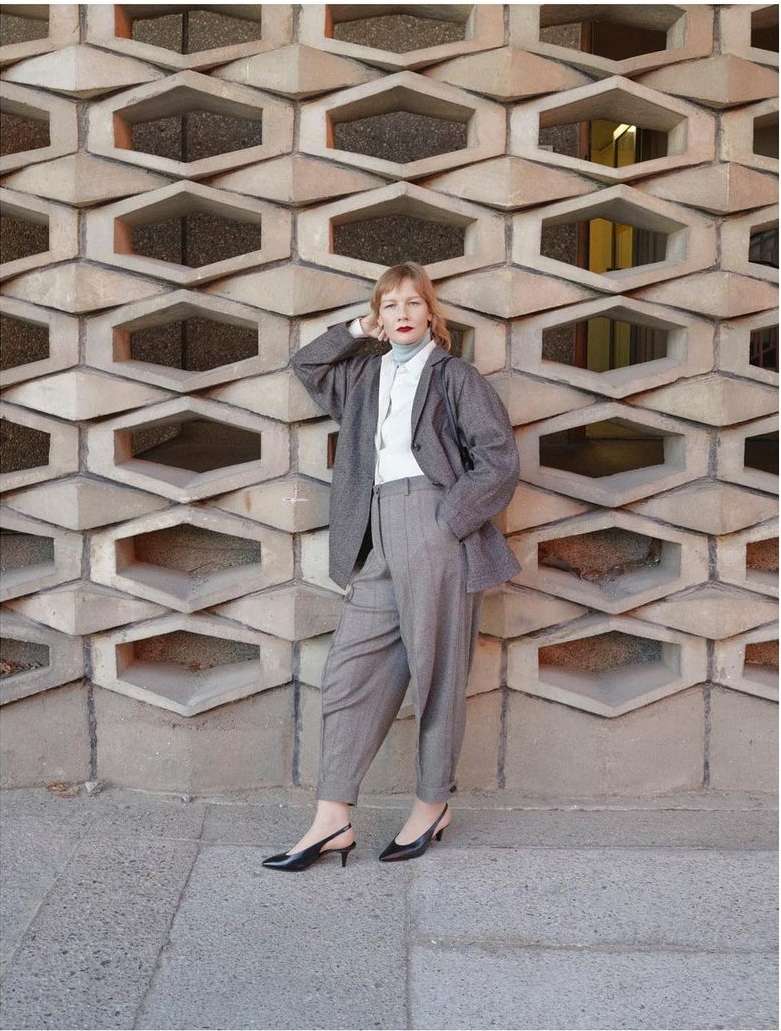 A woman in a grey suit leans against a honeycomb-patterned wall outside. She wears black slingback heels and a light blue turtleneck.