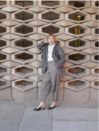 A woman in a grey suit leans against a honeycomb-patterned wall outside. She wears black slingback heels and a light blue turtleneck.