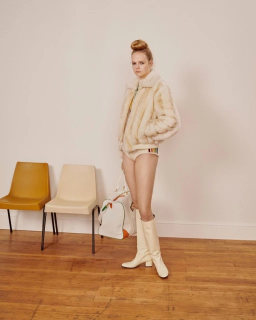 A young adult woman stands in a photo studio wearing a fur jacket, briefs, and boots, carrying a duffel bag in a medium shot.