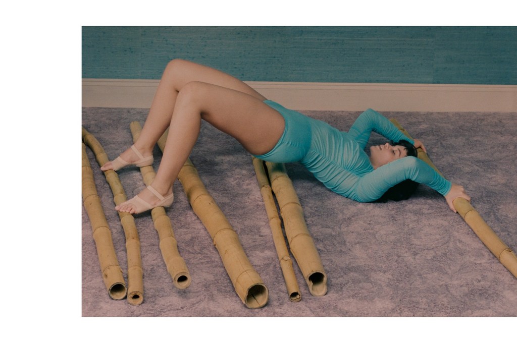 Woman in a blue bodysuit does a bridge pose on the floor, supported by bamboo sticks. Low angle, indoor environment, neutral color palette.