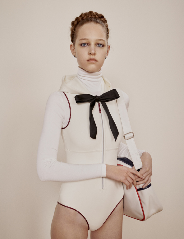 Young woman in cream bodysuit and turtle neck poses in front of a neutral wall with a bow and white sport tote bag.