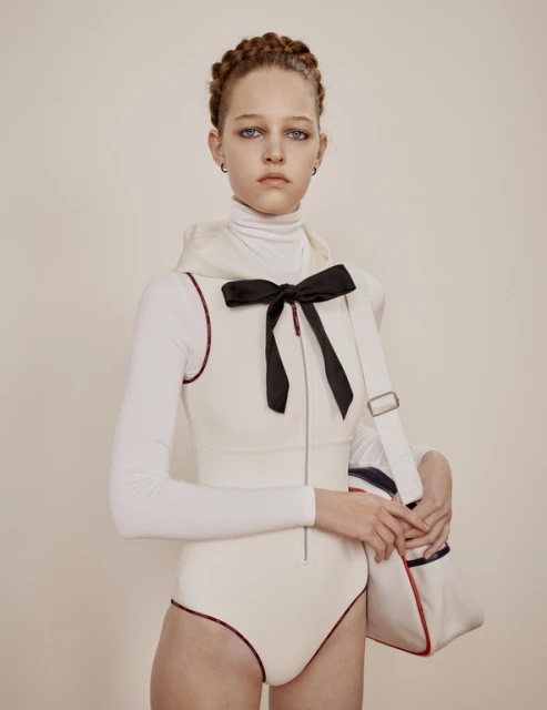 Young woman in cream bodysuit and turtle neck poses in front of a neutral wall with a bow and white sport tote bag.
